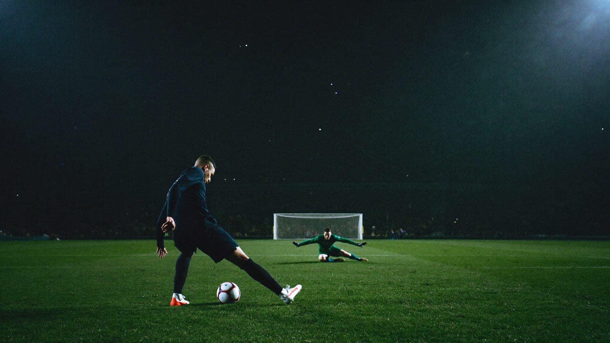 A footballer striking a penalty at night under stadium floodlights while the goalkeeper waits on the line